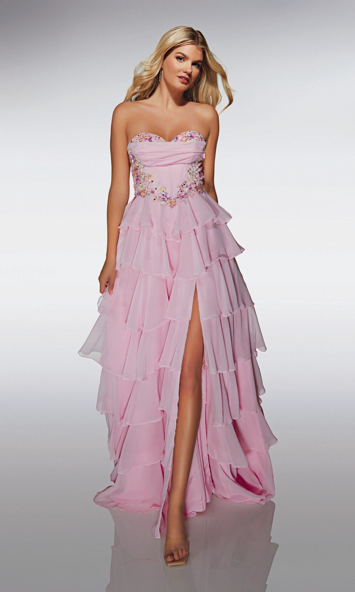 A woman wears the Alyce 62013 long prom dress in pink, featuring a strapless chiffon design with floral appliques, corset bodice, tiered skirt, and thigh-high slit, standing against a plain background.